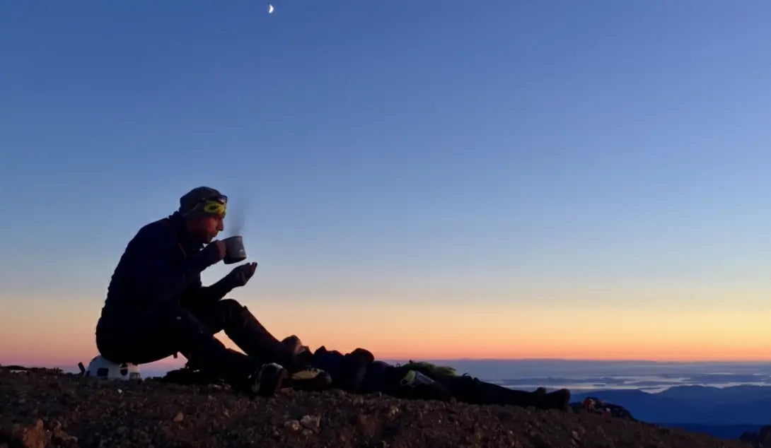 Person sitting on a mountain top at sunset or sunrise, holding a mug.